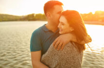 A couple embraces near a serene lake during sunset. The man, wearing a blue polo shirt, gently kisses the woman's forehead. She smiles, wearing a gray sweater, and her hair blows gently in the breeze. Warm sunlight casts a golden glow over them.