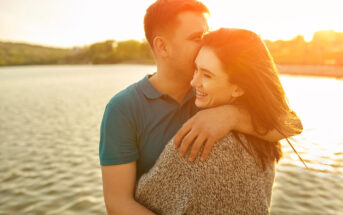 A couple embraces near a serene lake during sunset. The man, wearing a blue polo shirt, gently kisses the woman's forehead. She smiles, wearing a gray sweater, and her hair blows gently in the breeze. Warm sunlight casts a golden glow over them.