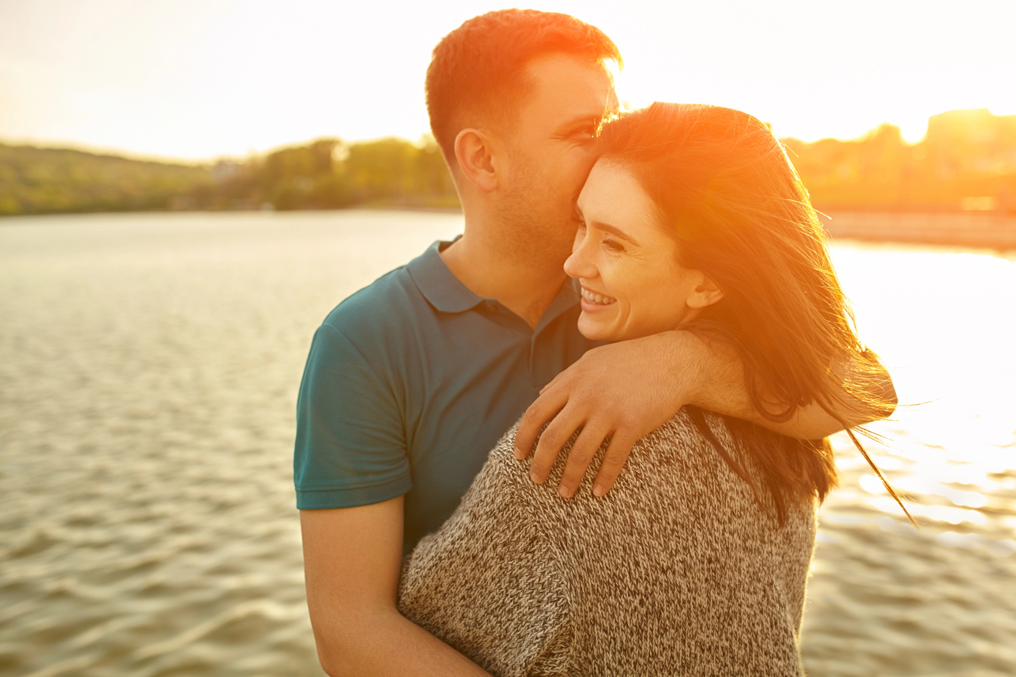 A couple embraces near a serene lake during sunset. The man, wearing a blue polo shirt, gently kisses the woman's forehead. She smiles, wearing a gray sweater, and her hair blows gently in the breeze. Warm sunlight casts a golden glow over them.