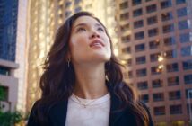 A woman with long, wavy hair gazes upwards, standing amidst tall, modern office buildings. She wears a white shirt and a black jacket. The background shows the sun reflecting off the glass and metal surfaces of the buildings, adding a warm glow to the urban scene.