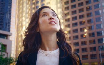 A woman with long, wavy hair gazes upwards, standing amidst tall, modern office buildings. She wears a white shirt and a black jacket. The background shows the sun reflecting off the glass and metal surfaces of the buildings, adding a warm glow to the urban scene.