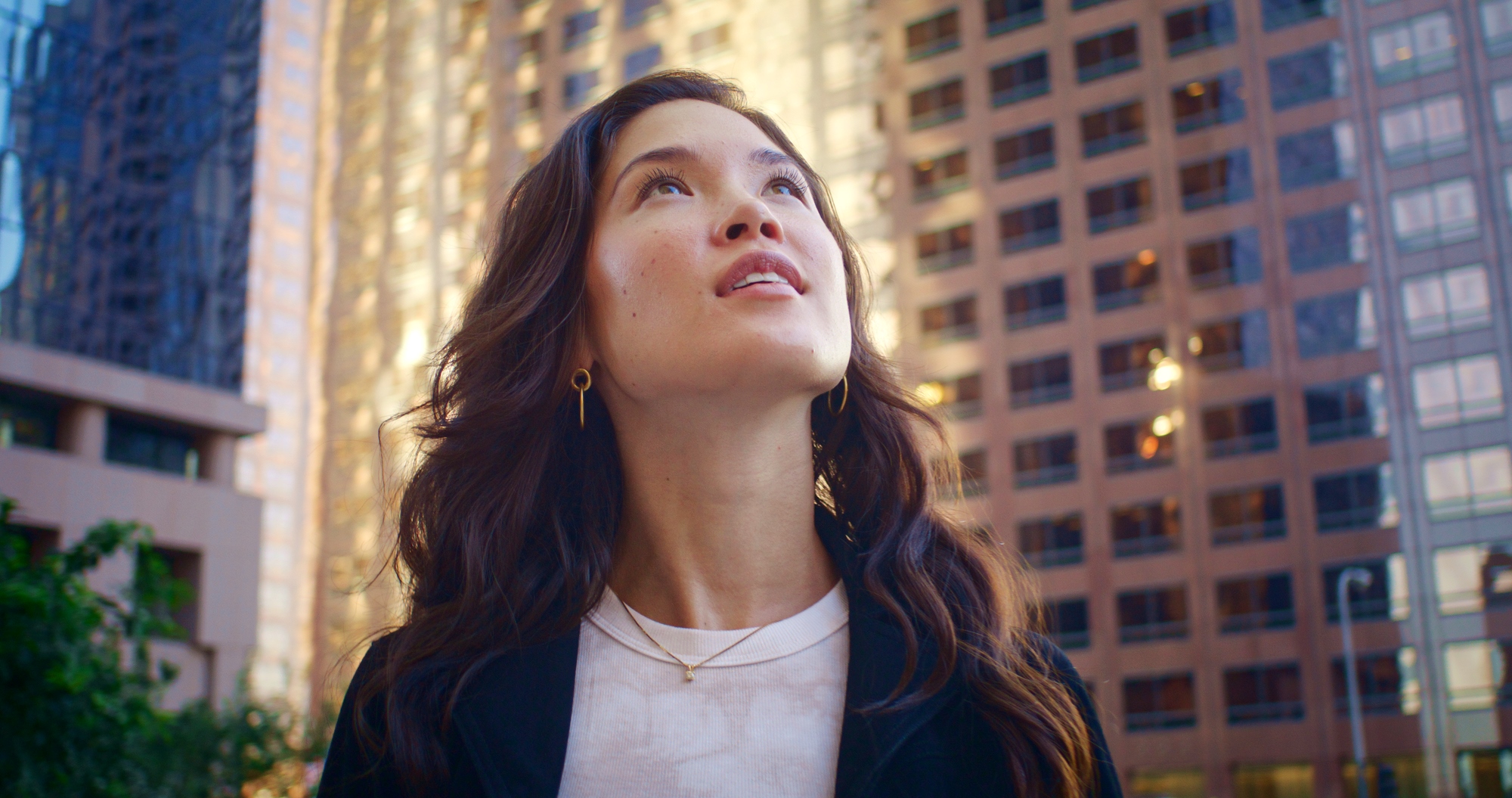 A woman with long, wavy hair gazes upwards, standing amidst tall, modern office buildings. She wears a white shirt and a black jacket. The background shows the sun reflecting off the glass and metal surfaces of the buildings, adding a warm glow to the urban scene.