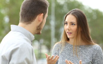 Two people are engaged in a conversation outdoors. The woman on the right, with long brown hair and wearing a gray sweater, is gesturing with her hands and has an expressive look on her face. The man on the left, with short brown hair, faces away from the camera.