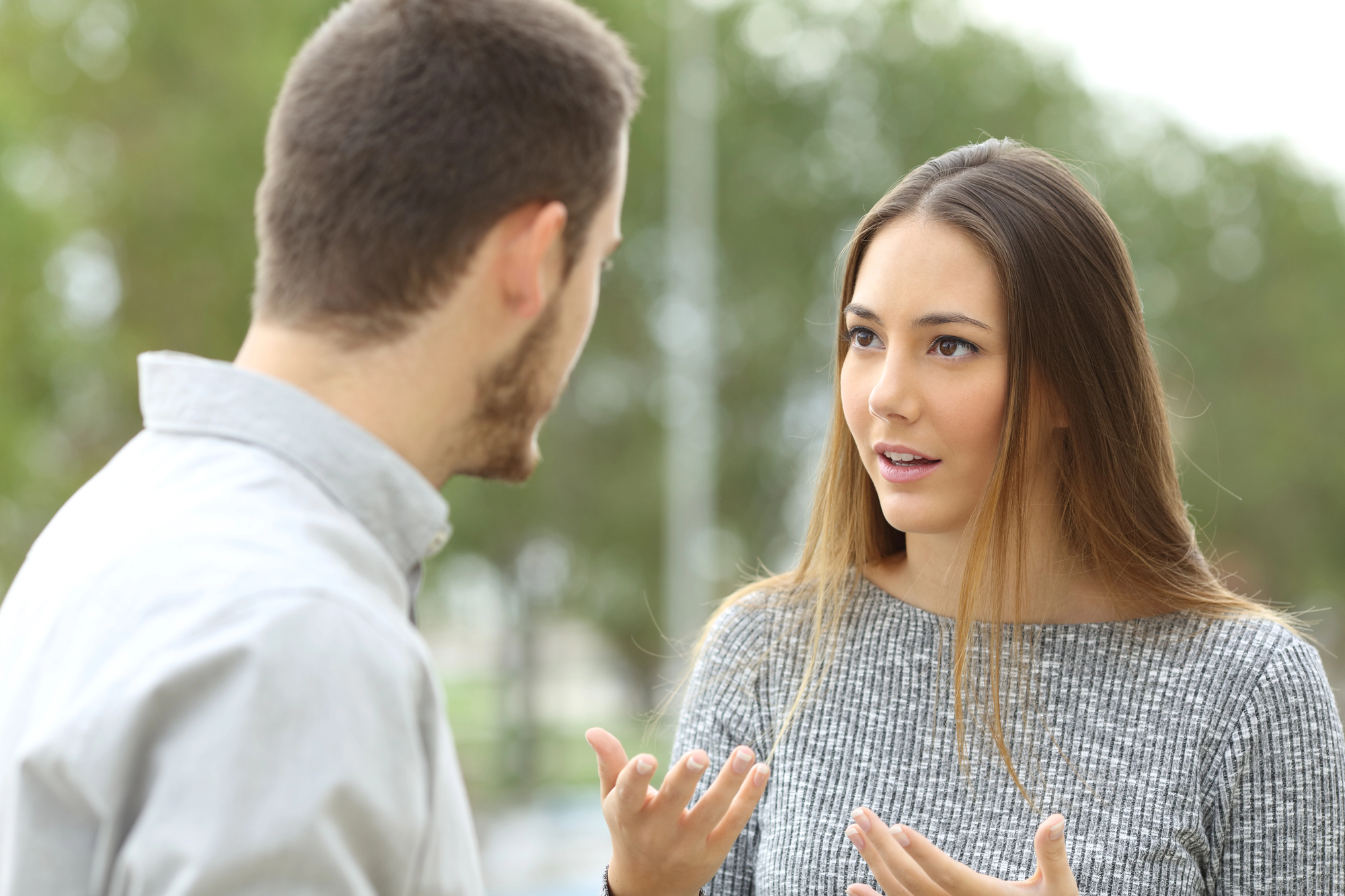 Two people are engaged in a conversation outdoors. The woman on the right, with long brown hair and wearing a gray sweater, is gesturing with her hands and has an expressive look on her face. The man on the left, with short brown hair, faces away from the camera.