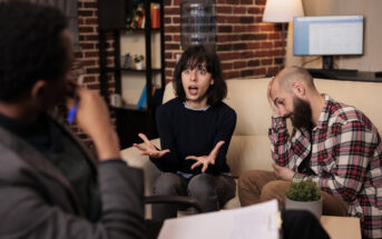 In a room with a brick wall, three people are engaged in an intense discussion. One person in the middle gestures animatedly with a surprised expression, while the person to the right has a distressed look, with their head resting on one hand. The third person listens attentively.