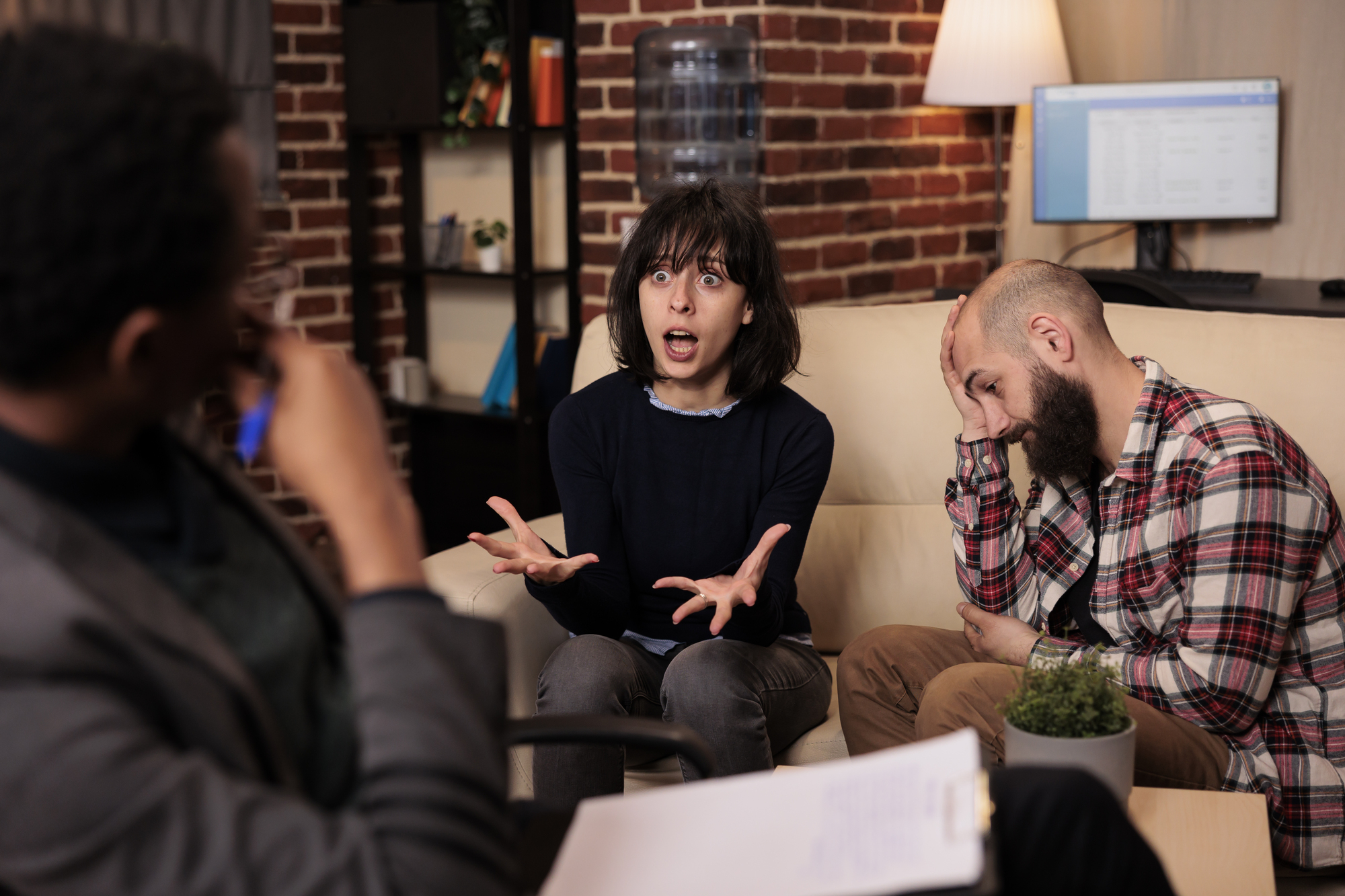 In a room with a brick wall, three people are engaged in an intense discussion. One person in the middle gestures animatedly with a surprised expression, while the person to the right has a distressed look, with their head resting on one hand. The third person listens attentively.