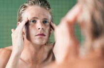 A person with light hair and blue eyes is standing in front of a mirror, gently touching their temples with both hands. They appear to be looking closely at their reflection, possibly inspecting their skin or face. The background has a green tiled wall.