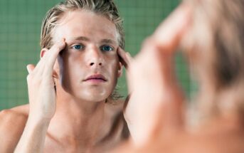 A person with light hair and blue eyes is standing in front of a mirror, gently touching their temples with both hands. They appear to be looking closely at their reflection, possibly inspecting their skin or face. The background has a green tiled wall.