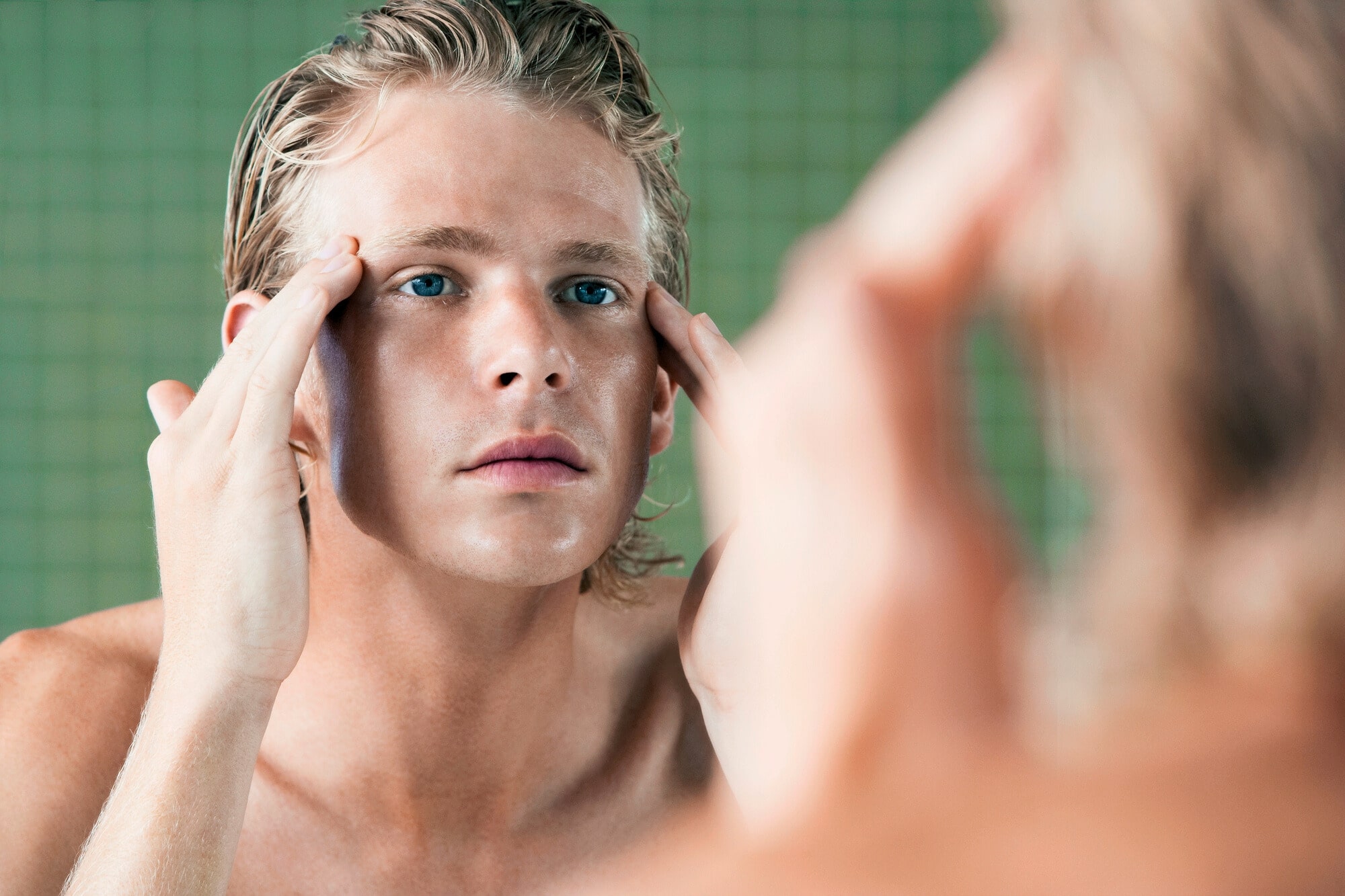 A person with light hair and blue eyes is standing in front of a mirror, gently touching their temples with both hands. They appear to be looking closely at their reflection, possibly inspecting their skin or face. The background has a green tiled wall.