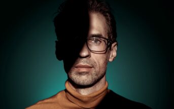 A man wearing glasses and a turtleneck sweater stands against a teal background with dramatic lighting. Half of his face is in shadow, creating a striking contrast between light and dark.