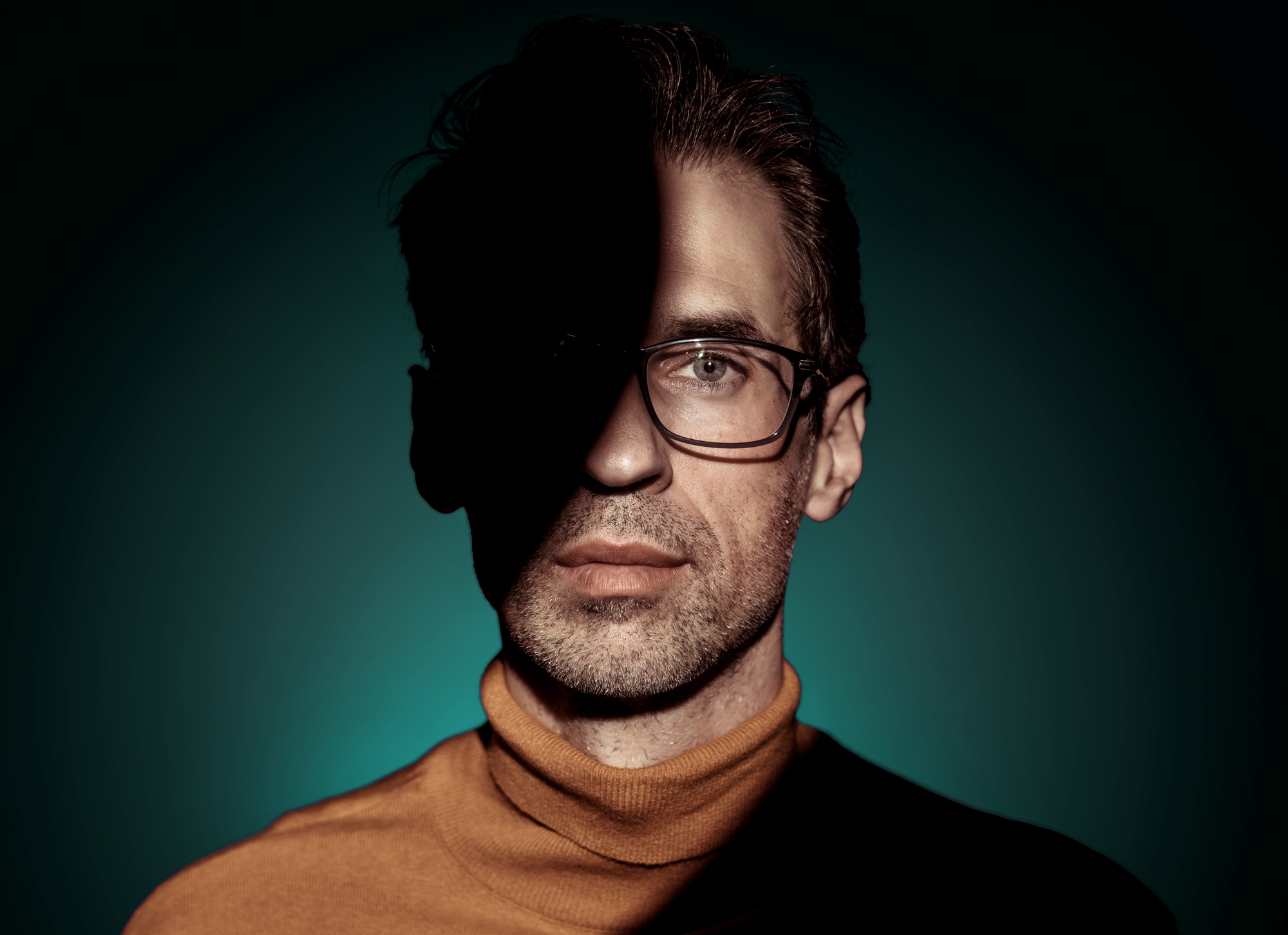 A man wearing glasses and a turtleneck sweater stands against a teal background with dramatic lighting. Half of his face is in shadow, creating a striking contrast between light and dark.