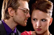 A man with glasses whispers closely to a woman with an anxious expression. The man has short, light hair and a beard, while the woman has her hair tied up and is wearing a red dress. The background is dark, highlighting their facial expressions.