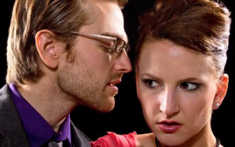 A man with glasses whispers closely to a woman with an anxious expression. The man has short, light hair and a beard, while the woman has her hair tied up and is wearing a red dress. The background is dark, highlighting their facial expressions.