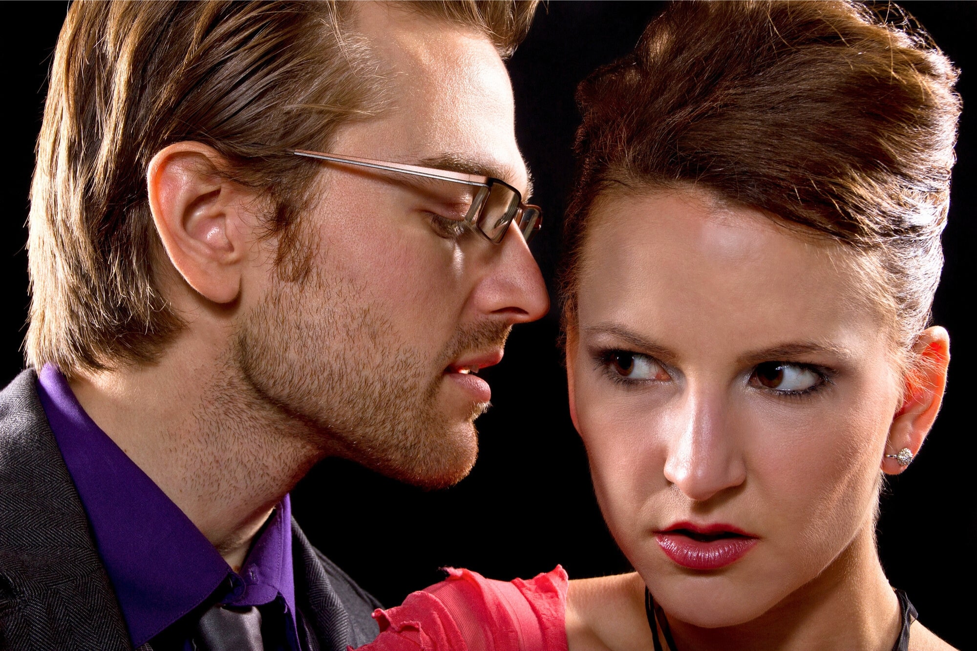 A man with glasses whispers closely to a woman with an anxious expression. The man has short, light hair and a beard, while the woman has her hair tied up and is wearing a red dress. The background is dark, highlighting their facial expressions.