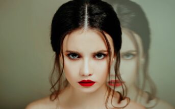 A woman with pale skin, dark hair, and bold red lipstick gazes intently at the camera. Her hair is neatly styled, and she has a serious expression. To the right, a slightly transparent reflection of her face adds a surreal effect to the image.