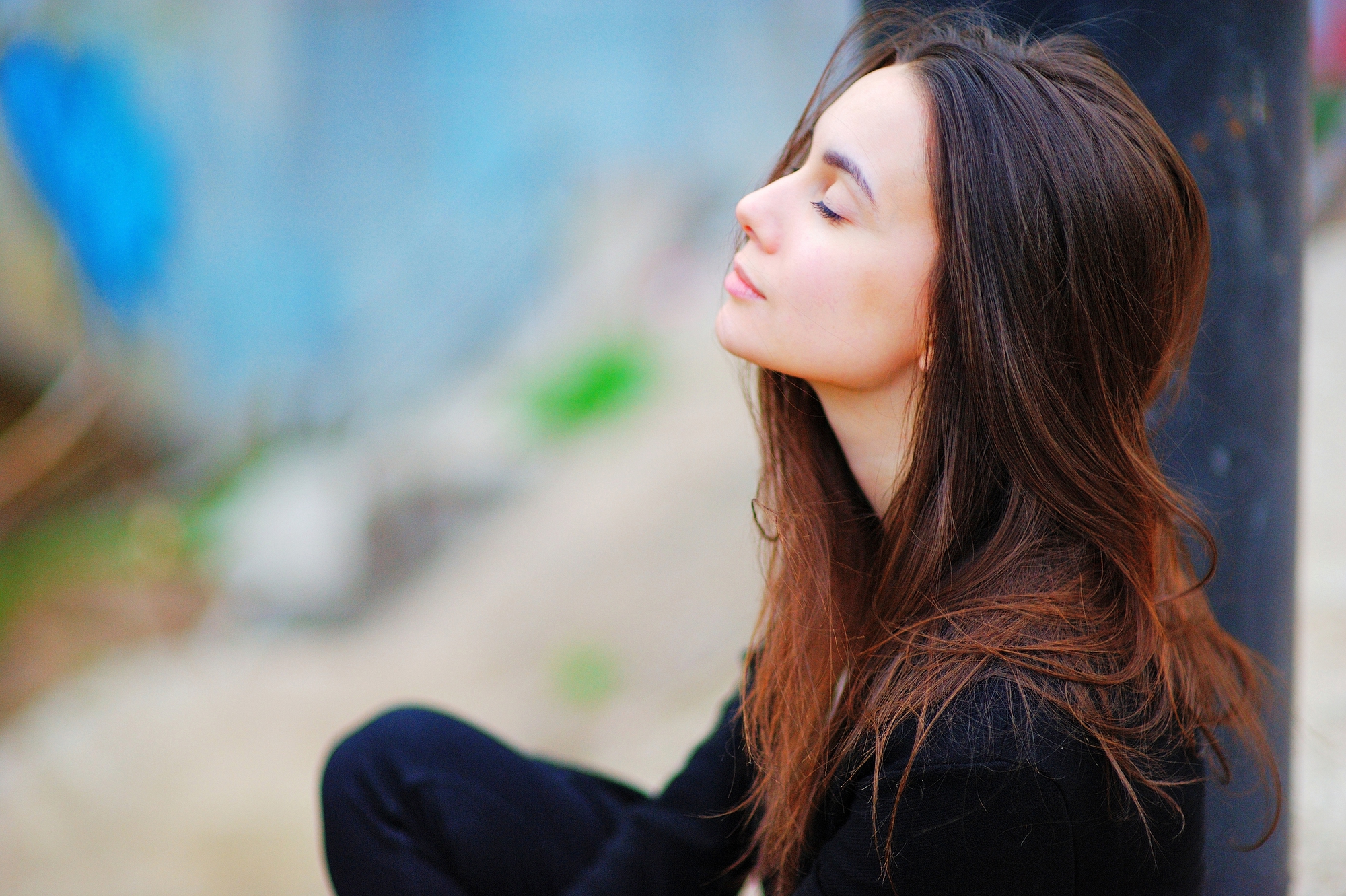 A young woman with long brown hair sits outdoors with her eyes closed in a peaceful expression. She is wearing a black top and leans against a dark pillar. The background is blurred, showing hints of greenery and urban elements.