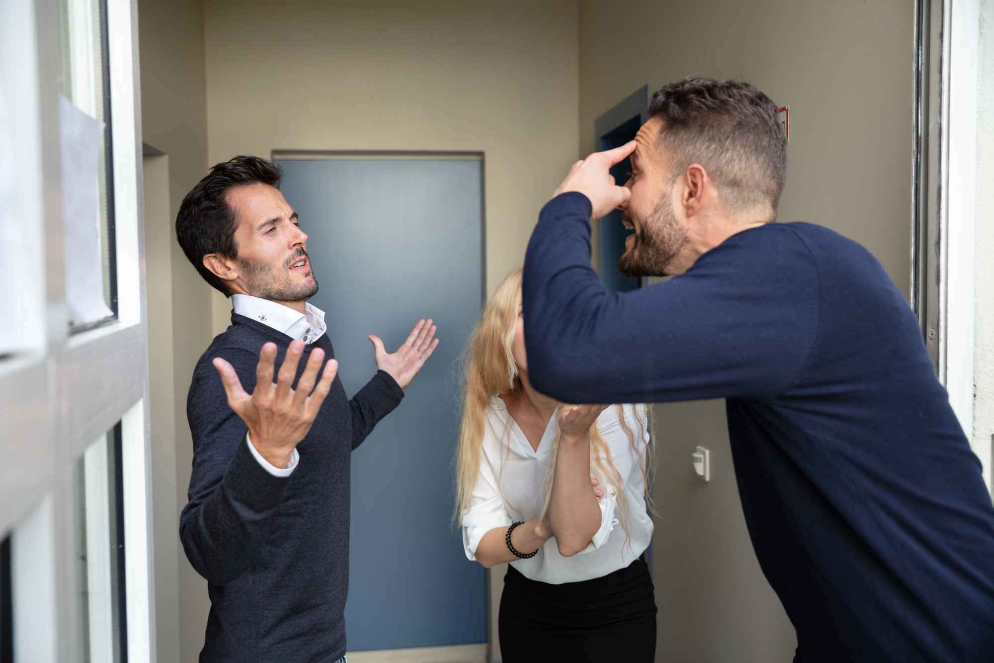 Three people are in a hallway, seemingly having a heated conversation. A man on the left has his arms raised and appears frustrated. Another man on the right is gesturing and looks upset. A woman in the middle has her head down and appears distressed.