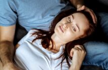 A woman with red hair wearing a white shirt lies on a person's lap, resting her head and looking upwards. The person she is resting on is wearing a blue shirt and gently touching her head. They both appear relaxed.