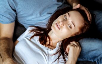 A woman with red hair wearing a white shirt lies on a person's lap, resting her head and looking upwards. The person she is resting on is wearing a blue shirt and gently touching her head. They both appear relaxed.