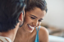 A close-up of a smiling woman with her hair tied back, leaning in toward a person with short, dark hair whose face is partially visible. The woman is wearing a light blue top and appears to be enjoying a pleasant moment with the other person.