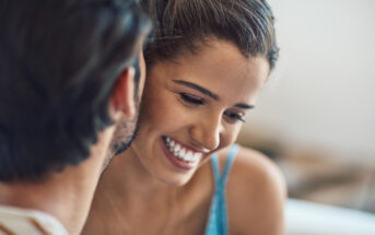 A close-up of a smiling woman with her hair tied back, leaning in toward a person with short, dark hair whose face is partially visible. The woman is wearing a light blue top and appears to be enjoying a pleasant moment with the other person.