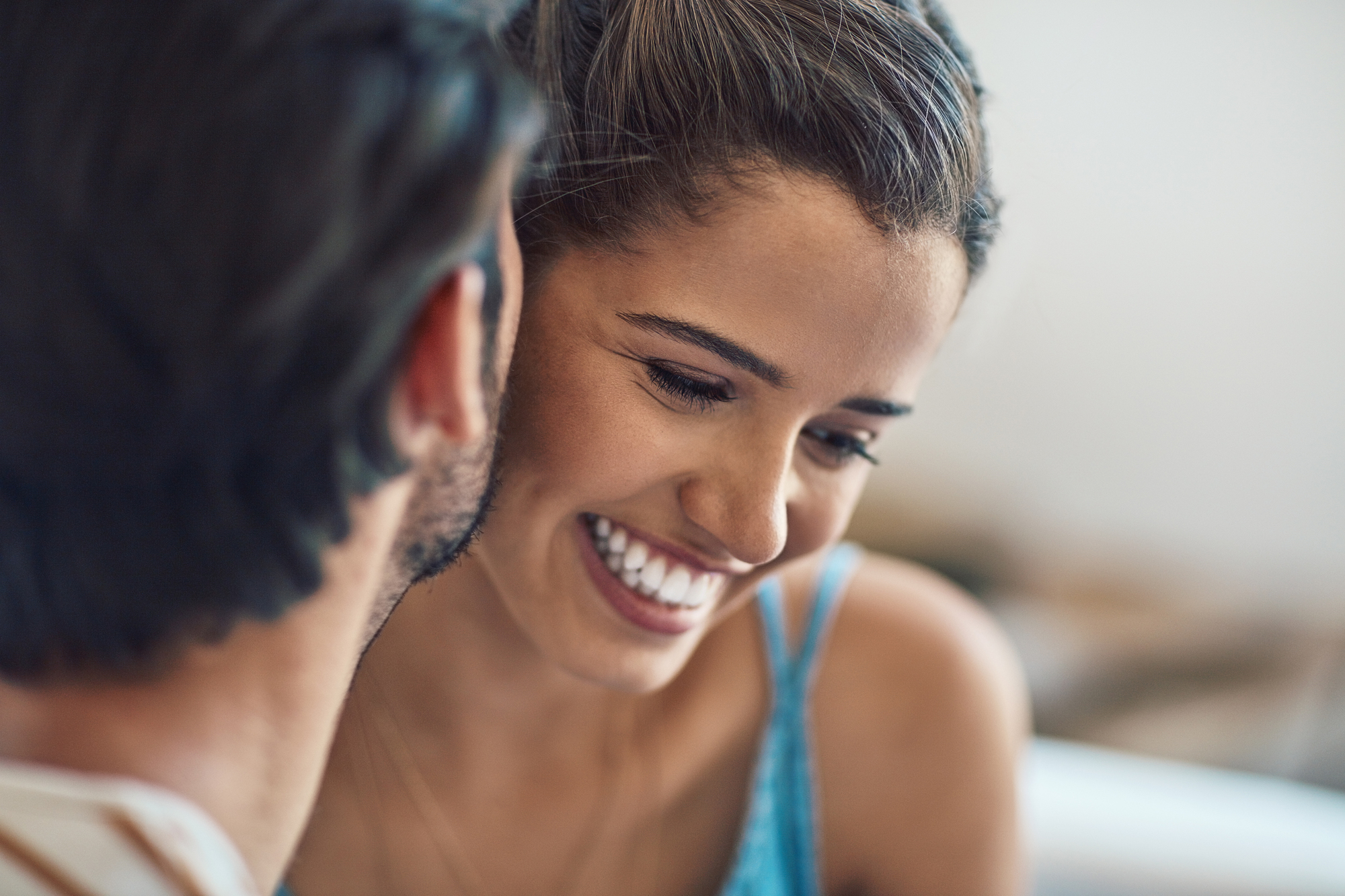 A close-up of a smiling woman with her hair tied back, leaning in toward a person with short, dark hair whose face is partially visible. The woman is wearing a light blue top and appears to be enjoying a pleasant moment with the other person.