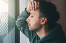 A man with short dark hair and a beard rests his hand on his forehead and leans against a window, eyes closed, appearing deep in thought. He is wearing a green hoodie, and light streams in through the window, illuminating part of his face.