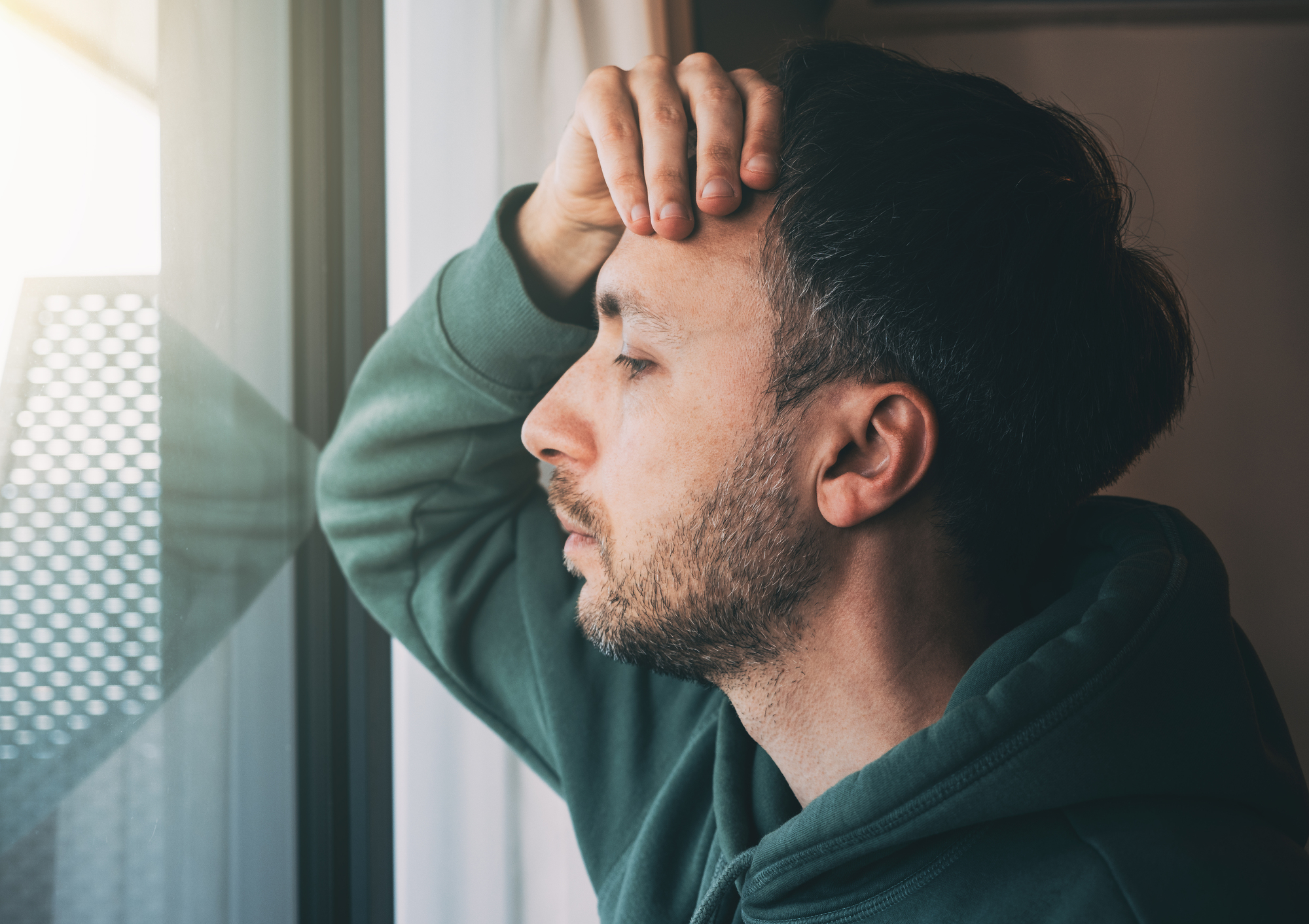 A man with short dark hair and a beard rests his hand on his forehead and leans against a window, eyes closed, appearing deep in thought. He is wearing a green hoodie, and light streams in through the window, illuminating part of his face.