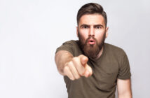 A man with a beard and short hair, wearing a green T-shirt, points towards the camera with an intense expression, as if surprised or accusing. He stands against a plain white background.