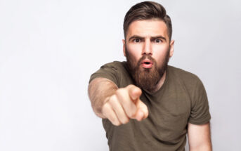 A man with a beard and short hair, wearing a green T-shirt, points towards the camera with an intense expression, as if surprised or accusing. He stands against a plain white background.