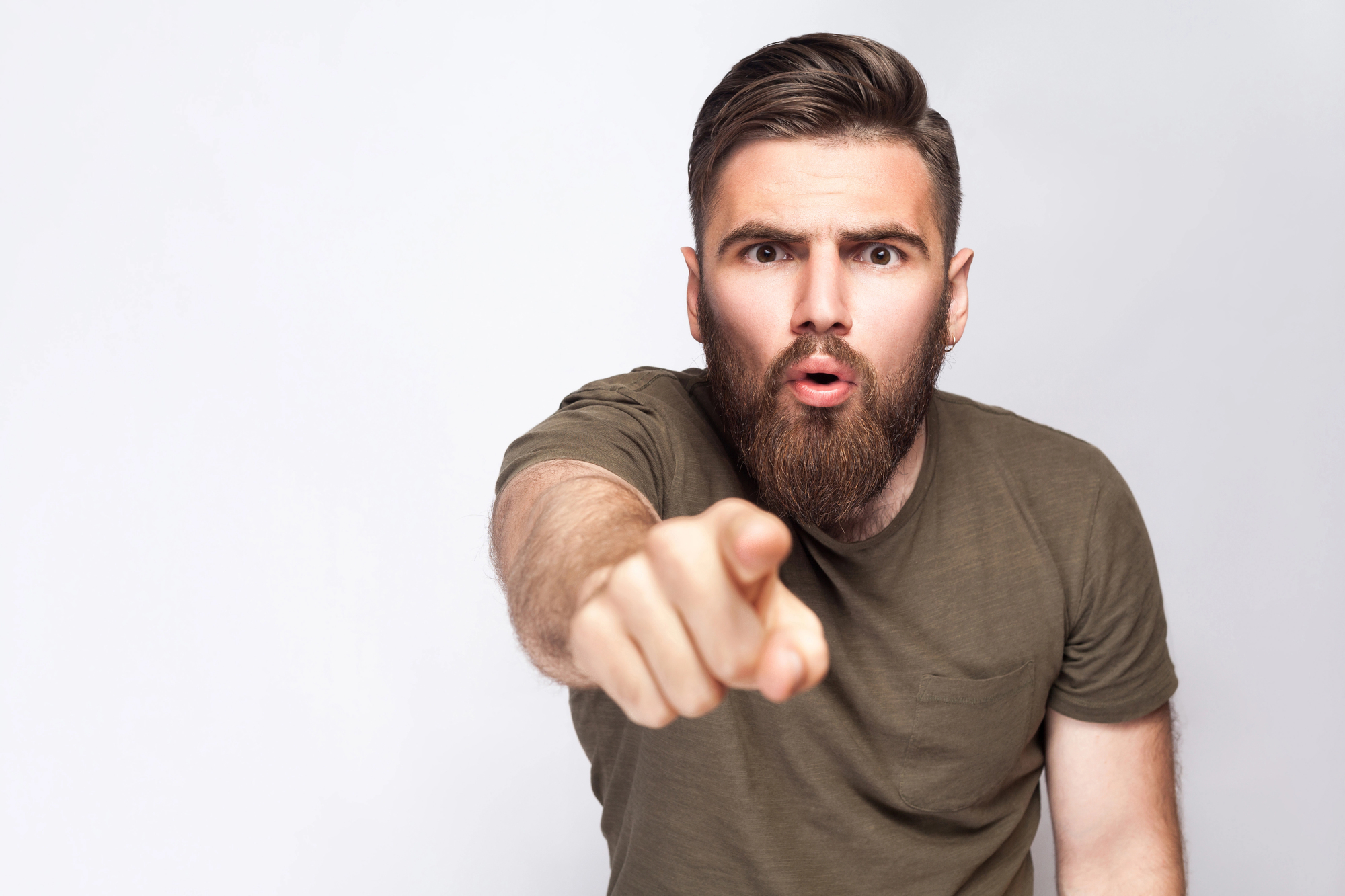 A man with a beard and short hair, wearing a green T-shirt, points towards the camera with an intense expression, as if surprised or accusing. He stands against a plain white background.