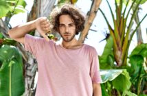 A young man with wavy hair wearing a pink t-shirt stands outdoors in front of tropical plants. He is looking at the camera with a neutral expression and giving a thumbs-down gesture with his right hand.