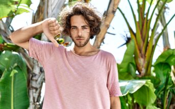 A young man with wavy hair wearing a pink t-shirt stands outdoors in front of tropical plants. He is looking at the camera with a neutral expression and giving a thumbs-down gesture with his right hand.