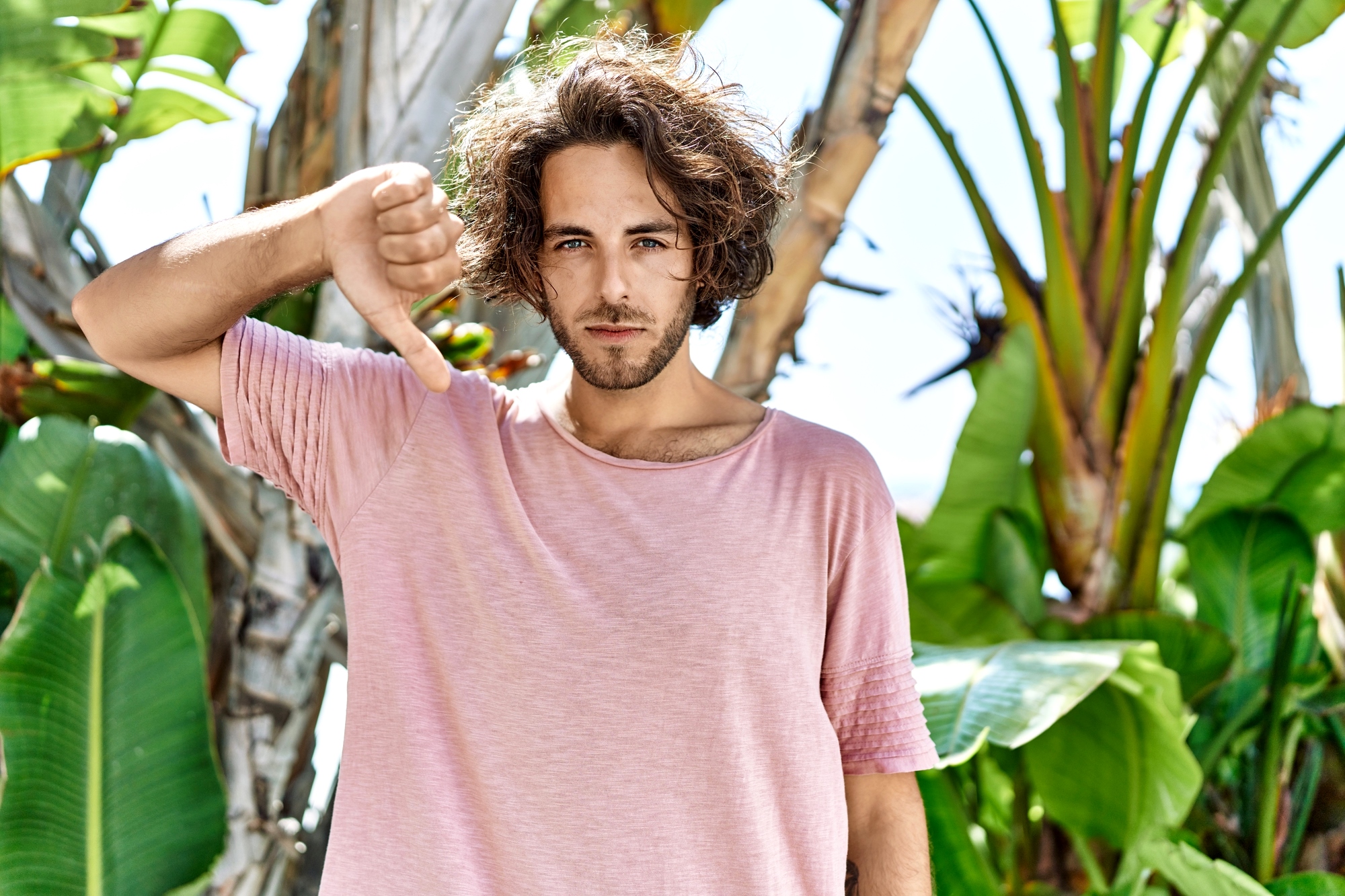 A young man with wavy hair wearing a pink t-shirt stands outdoors in front of tropical plants. He is looking at the camera with a neutral expression and giving a thumbs-down gesture with his right hand.