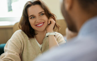 A woman with shoulder-length brown hair and a beige sweater smiles warmly while resting her head on her hand. She appears to be in conversation with a person in the foreground, who is partially visible and facing her. The background is softly blurred.