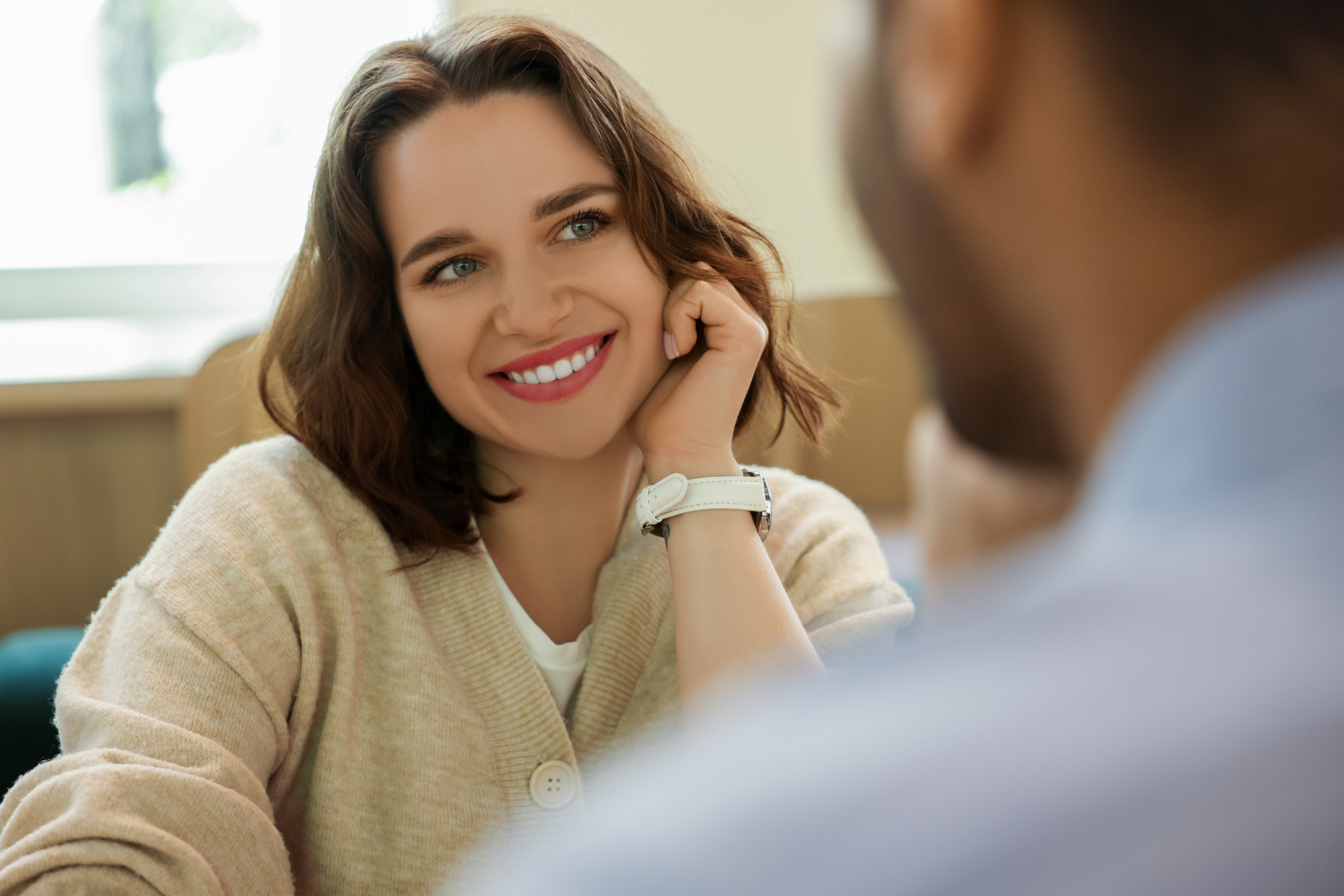 A woman with shoulder-length brown hair and a beige sweater smiles warmly while resting her head on her hand. She appears to be in conversation with a person in the foreground, who is partially visible and facing her. The background is softly blurred.