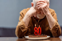 An elderly man with a sad expression sits at a table with his head in his hands. In front of him is a birthday cake with lit candles shaped as the number 80. He is wearing a brown shirt and has white hair. The background is blurred and grayish blue.