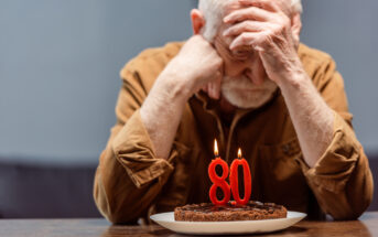 An elderly man with a sad expression sits at a table with his head in his hands. In front of him is a birthday cake with lit candles shaped as the number 80. He is wearing a brown shirt and has white hair. The background is blurred and grayish blue.