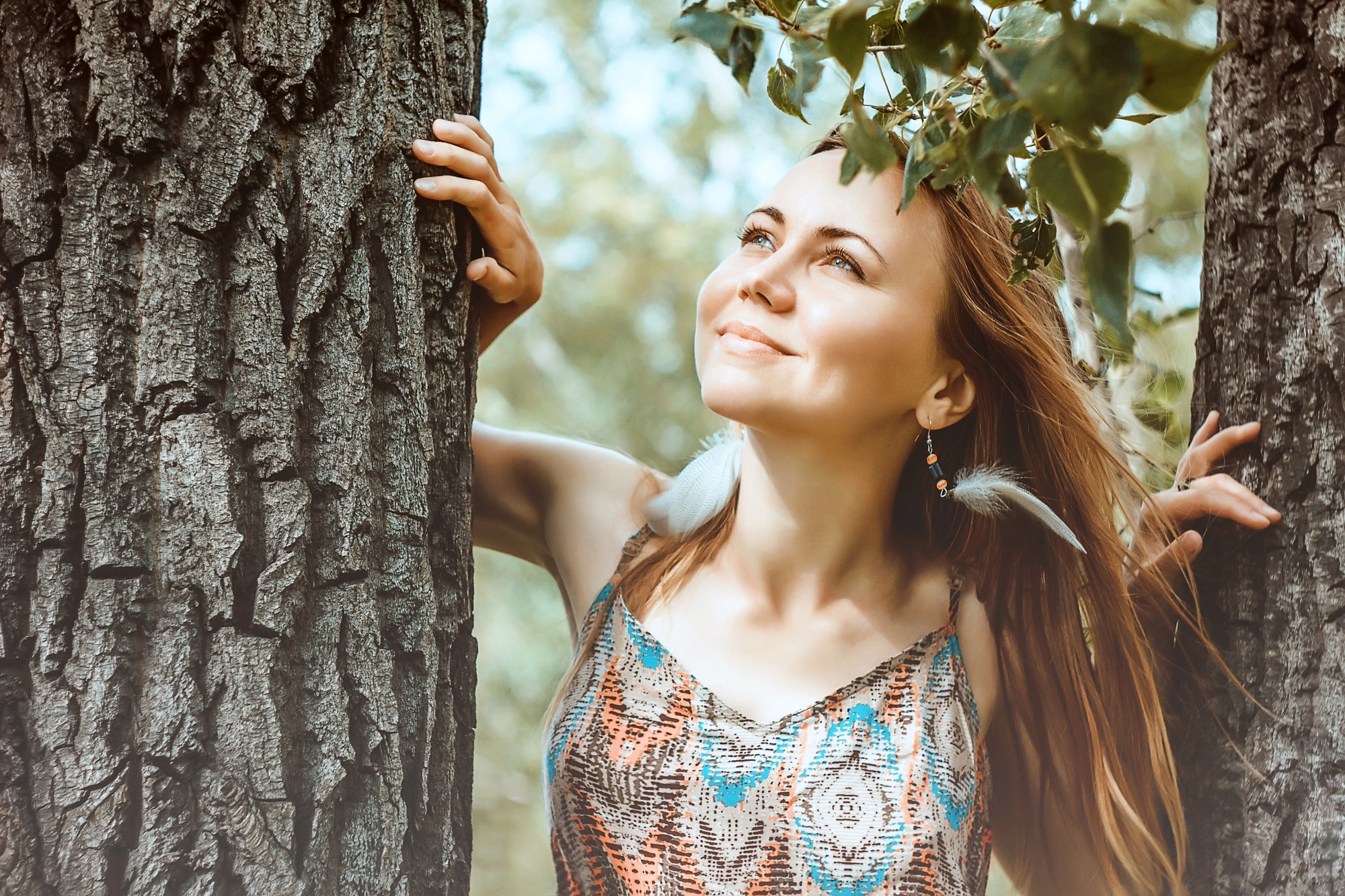 A woman with long hair and feather earrings stands between two trees, looking up with a smile. She is wearing a patterned sleeveless dress. Sunlight filters through green leaves above her, creating a serene, nature-filled atmosphere.