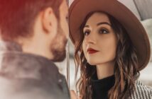 A woman with long curly hair, wearing a wide-brimmed hat and red lipstick, gazes at a man under an umbrella on a rainy day. The man has a beard and is facing her, with only the back of his head visible.