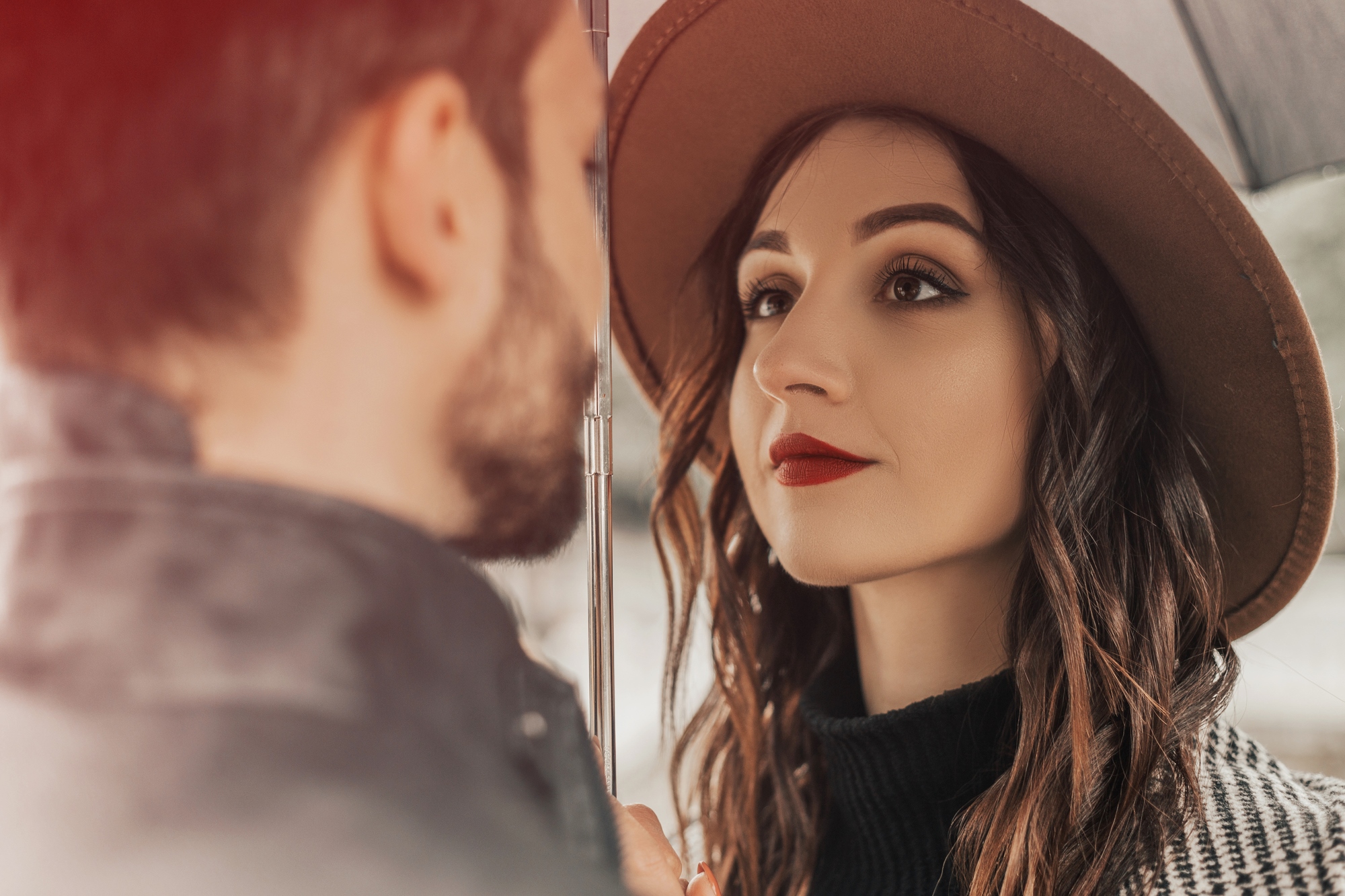 A woman with long curly hair, wearing a wide-brimmed hat and red lipstick, gazes at a man under an umbrella on a rainy day. The man has a beard and is facing her, with only the back of his head visible.