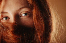 Close-up of a person with green eyes and long red hair partially covering their face. The light highlights the texture and color of their hair and the detail in their eyes. The background is a warm, earthy tone, complementing the overall warm hues of the image.