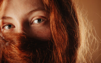 Close-up of a person with green eyes and long red hair partially covering their face. The light highlights the texture and color of their hair and the detail in their eyes. The background is a warm, earthy tone, complementing the overall warm hues of the image.