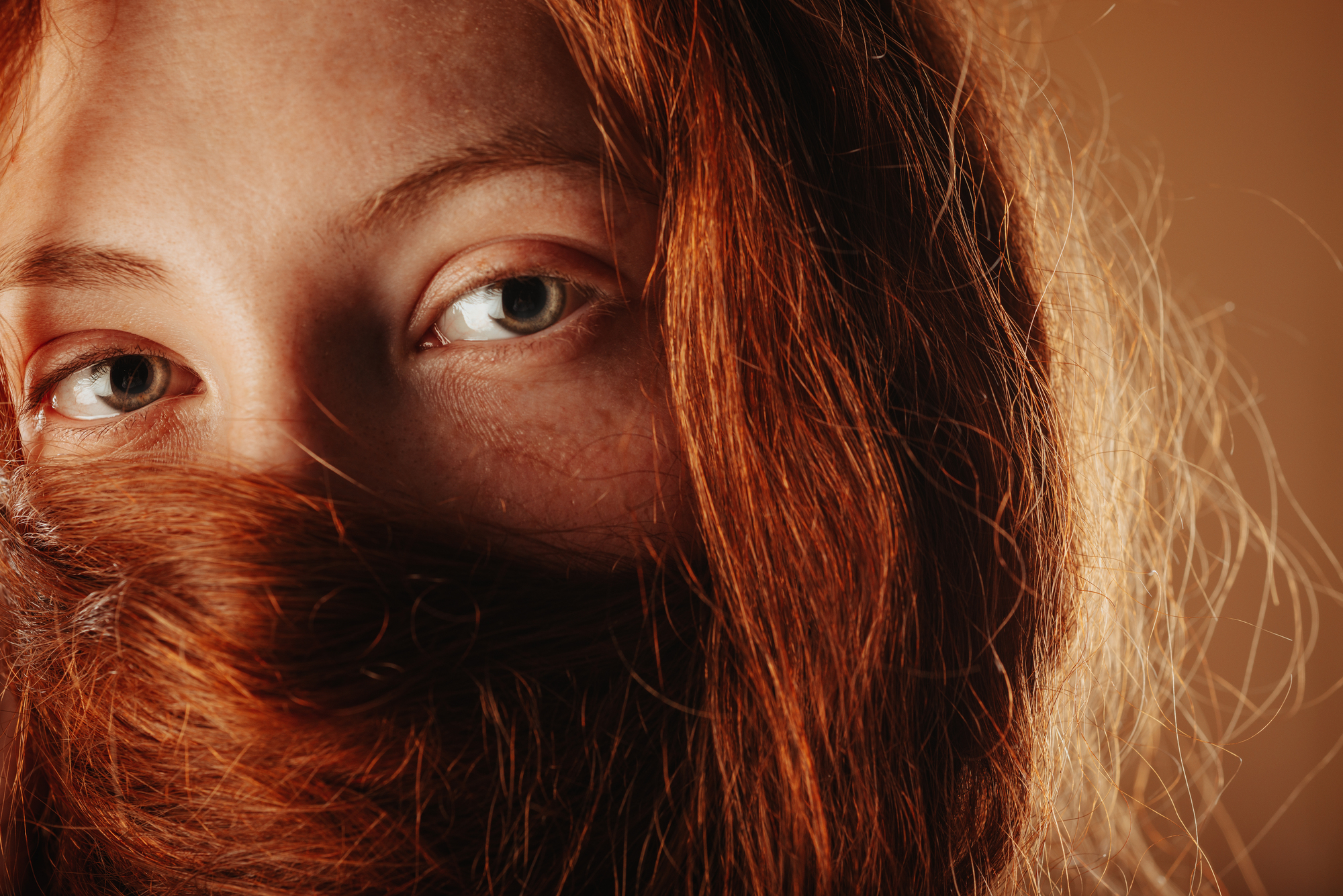 Close-up of a person with green eyes and long red hair partially covering their face. The light highlights the texture and color of their hair and the detail in their eyes. The background is a warm, earthy tone, complementing the overall warm hues of the image.