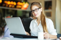 A person with long hair, wearing glasses and a white shirt, sits at a table using a laptop. They are holding a white mug in their left hand and are in a modern, well-lit cafe environment with blurred customers and menu boards in the background.