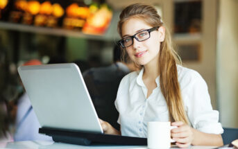 A person with long hair, wearing glasses and a white shirt, sits at a table using a laptop. They are holding a white mug in their left hand and are in a modern, well-lit cafe environment with blurred customers and menu boards in the background.