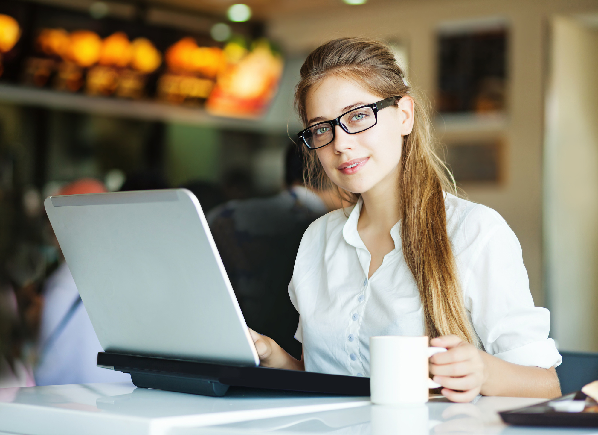 A person with long hair, wearing glasses and a white shirt, sits at a table using a laptop. They are holding a white mug in their left hand and are in a modern, well-lit cafe environment with blurred customers and menu boards in the background.