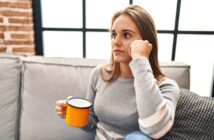 A woman with long brown hair sits on a couch, holding a yellow mug in her hand. She gazes thoughtfully out of a window with a pensive expression. She is wearing a grey sweater with white stripes on the sleeves. The background shows a brick wall and large window panes.