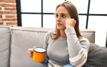 A woman with long brown hair sits on a couch, holding a yellow mug in her hand. She gazes thoughtfully out of a window with a pensive expression. She is wearing a grey sweater with white stripes on the sleeves. The background shows a brick wall and large window panes.