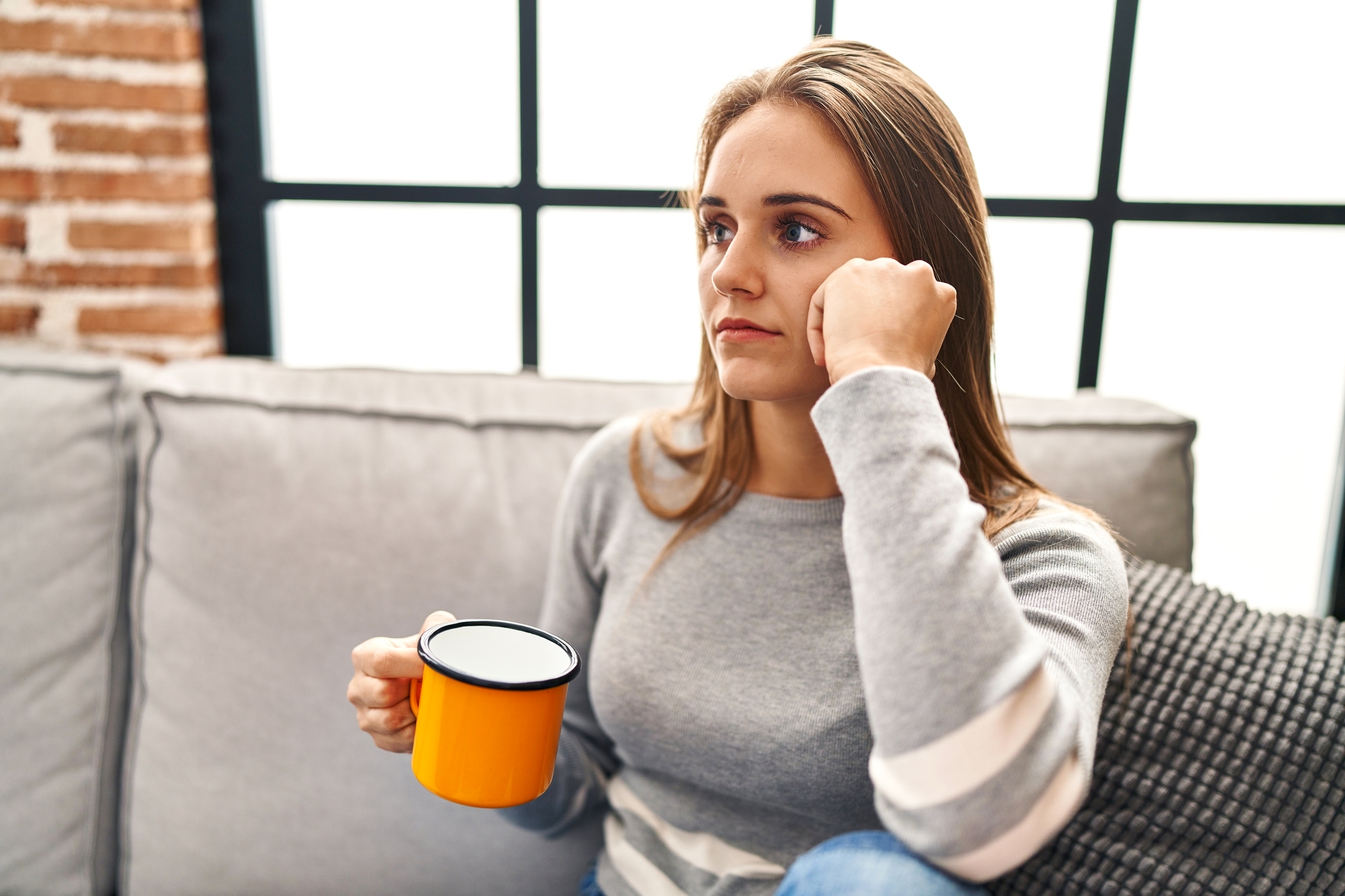 A woman with long brown hair sits on a couch, holding a yellow mug in her hand. She gazes thoughtfully out of a window with a pensive expression. She is wearing a grey sweater with white stripes on the sleeves. The background shows a brick wall and large window panes.